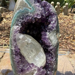 May include: A large amethyst geode with a quartz crystal formation inside. The geode has a dark purple interior with crystal formations and a gray, green, and white exterior. The quartz crystal is clear and elongated.