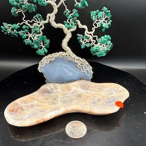 May include: A handcrafted wire tree sculpture with green malachite leaves sits atop a blue agate base. A beige and brown stone dish and a quarter are in the foreground. The dish has an orange price tag.