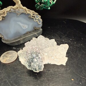 May include: A quarter coin next to a cluster of clear and purple crystals. A light blue and gray stone is in the background, with a silver wire tree and green malachite leaves. The objects are on a dark surface.