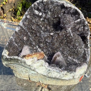 May include: A large, hollow amethyst geode featuring deep purple, crystalline structures lining the interior cavity. The exterior displays a rough, greyish-brown rock formation with a distinct, smooth, light-colored base and a small orange inclusion.