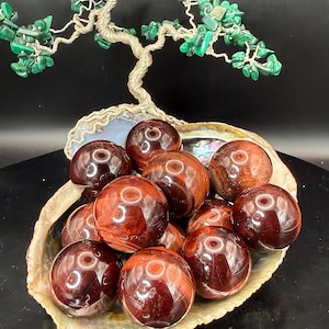 May include: A collection of polished, round, red tiger eye gemstone spheres displayed in a large, iridescent seashell. A decorative tree sculpture with green malachite leaves and a silver wire trunk sits above the shell.
