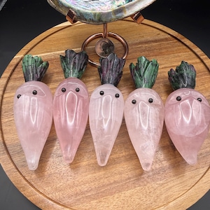 May include: Five pink quartz carrot figurines with carved green tops and small black eyes are arranged on a round wooden tray. The tray sits beneath a decorative iridescent shell.