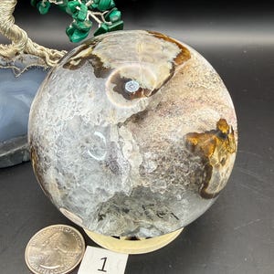 May include: A large, polished crystal sphere with a mix of clear, brown, and white tones. The sphere sits on a light-colored stand. A US quarter coin is next to the sphere, and a small white tag with the number "1" is visible.