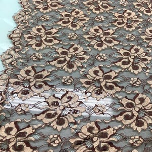 Può includere: Primo piano di un tessuto di pizzo floreale. Il pizzo presenta un motivo ripetuto di grandi fiori stilizzati in tonalità marroni e beige su uno sfondo marrone scuro. Il design delicato è visibile in tutto il tessuto.