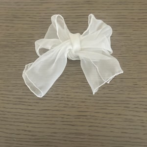 Puede incluir: Un delicado lazo blanco de gasa, probablemente para el cabello o envolver regalos, está centrado en una superficie de madera. El lazo está atado en el centro con extremos largos y fluidos. La tela parece ligera y aireada.