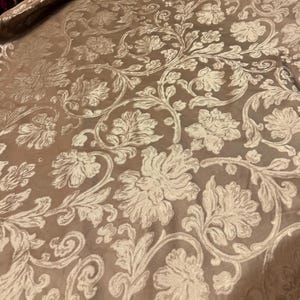 May include: Close-up of a fabric with a floral pattern. The fabric has a tan background with a cream-coloured floral design. The design includes large flowers, leaves, and swirling vines. The fabric appears to be soft and luxurious.
