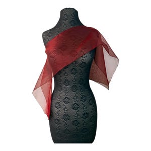 Peut inclure: Un châle en organza rouge foncé transparent drapé sur un mannequin noir au motif floral texturé. Le tissu léger et translucide crée des plis et des superpositions doux sur le haut du torse, s'étendant vers l'extérieur en une forme fluide.
