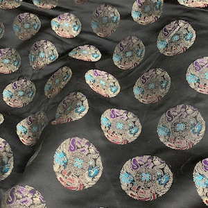 May include: Black fabric with an all-over pattern of stylized, oval-shaped medallions. Each medallion features a central geometric design, surrounded by dragons and floral elements in silver, purple, blue, and red. The fabric has a subtle sheen.