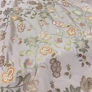 May include: Pale pink fabric with an embroidered floral pattern. The design features detailed flowers in shades of cream, gold, and muted green, with accents of red and brown. The fabric has a subtle sheen.