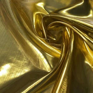 May include: A piece of metallic gold fabric with a high-gloss, reflective finish. The material is draped in soft, flowing folds, creating deep shadows and bright highlights across the surface that emphasize its smooth, shimmering texture.