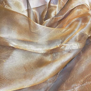 May include: Close-up of a shimmering, sheer fabric in shades of gold and bronze. The material is draped and folded, creating a textured, flowing effect. The fabric appears lightweight and delicate, with a subtle sheen.