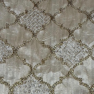 May include: Close-up of a cream-colored fabric with an intricate, repeating pattern. The design features a lattice-like structure with gold-colored outlines and a shimmering, textured background. The pattern is symmetrical and ornate.