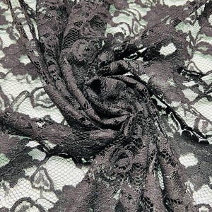 May include: Close-up of dark gray lace fabric with a floral pattern. The fabric is bunched and folded, showing the intricate details of the design. The lace has a delicate, open weave, creating a textured appearance.