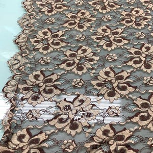 May include: Close-up of a floral lace fabric. The lace features a repeating pattern of large, stylized flowers in shades of brown and beige, set against a dark background. The delicate design is visible throughout the fabric.
