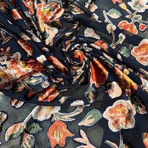 Può includere: Primo piano di un tessuto blu navy scuro con un motivo floreale. I fiori sono nelle tonalità dell'arancione, pesca e crema, con accenti verdi e blu. Il tessuto è leggermente traslucido, con una consistenza vellutata.