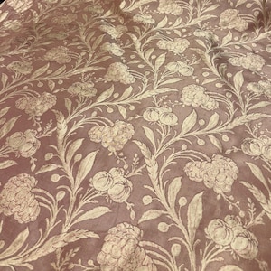 May include: A close-up of a dusty rose fabric with a repeating floral pattern. The design features cream-colored flowers, leaves, and vines. The fabric has a subtle sheen, suggesting a luxurious material.