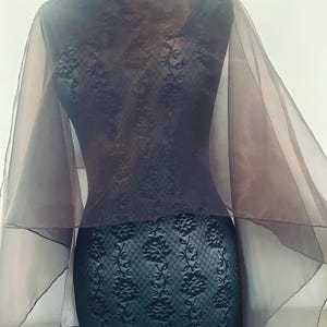 May include: A dark brown, semi-sheer organza capelet featuring a delicate floral lace pattern. The garment has a wide, boat-neck style neckline and flowing, translucent sleeves that drape over the shoulders, creating a layered appearance.