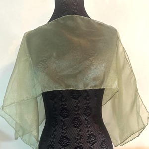 May include: A sheer, olive green shawl draped over a black mannequin. The fabric is lightweight and slightly textured, with a subtle sheen. The shawl is designed to be worn over the shoulders, with a rectangular shape and flared edges.