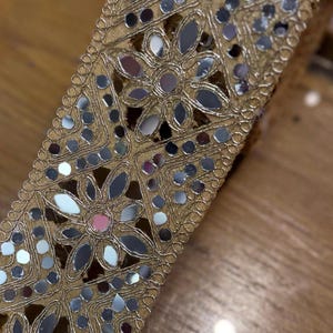 May include: Close-up of a decorative gold trim with floral and geometric patterns. The trim is embellished with small, reflective mirror pieces, creating a shimmering effect. The trim is likely used for embellishing clothing or crafts.