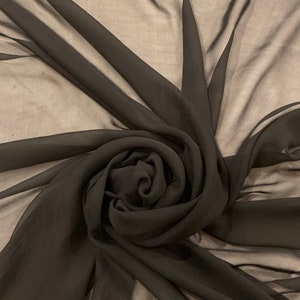 May include: A dark charcoal-colored fabric is artfully arranged in a spiral, creating a layered, textured effect. The sheer material allows light to filter through, highlighting the folds and curves of the fabric. The overall effect is elegant and flowing.