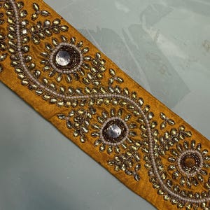 May include: Mustard yellow fabric trim with intricate embellishments. The trim features a repeating pattern of floral designs with clear and gold-colored beads and sequins. The design is bordered by small white beads.