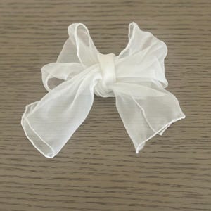Puede incluir: Un lazo de tela blanca y transparente, posiblemente para un accesorio para el cabello o un elemento decorativo. El lazo está atado en el centro con una pequeña banda, y la tela tiene una apariencia suave y fluida. El fondo es una superficie de madera marrón claro.