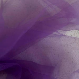 May include: Close-up of a piece of sheer, purple tulle fabric. The fabric is layered and has a delicate, net-like texture. The color varies in intensity, with deeper shades of purple in the folds and lighter hues in the open areas.