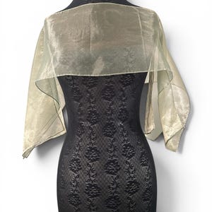May include: A sheer, light gold shawl with a subtle, tonal floral pattern draped over a black, textured, sleeveless garment. The shawl features a rectangular shape with finished edges, resting across the shoulders and upper chest area.