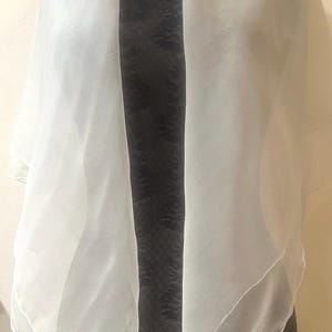 May include: A sheer white scarf draped over a black mannequin. The scarf is made of a lightweight, flowing fabric, and it is folded to create a layered effect. The scarf's edges are slightly rounded.