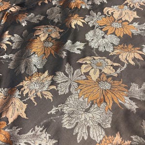 May include: Close-up of a fabric with a floral pattern. The design features large flowers in shades of gold, tan, and silver, set against a dark brown background. The leaves and petals have intricate details.