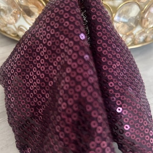 May include: Close-up of a deep purple sequined fabric. The fabric is folded, revealing the texture and sheen of the sequins. The sequins are circular and densely packed, creating a shimmering effect. The fabric is likely used for apparel or decorative purposes.
