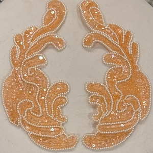 May include: Two beige sequined fabric appliques with a pearl-like trim. The appliques are shaped like a stylized floral design.