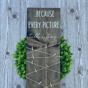 May include: A dark brown wooden sign with the text "... BECAUSE EVERY PICTURE tells a story..." in white lettering. The sign has twine strung across it with clothespins attached.