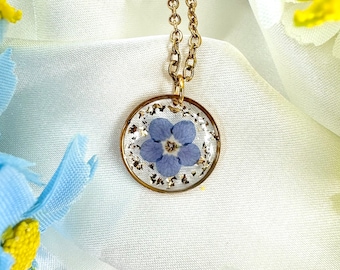 Puede incluir: Un collar de cadena dorada con un colgante circular. El colgante presenta una flor azul claro encapsulada en resina transparente, con copos dorados. El collar se muestra sobre un fondo de tela blanca.
