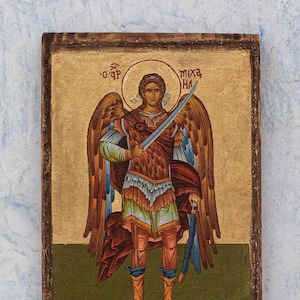 Puede incluir: Icono rectangular de madera que representa a un &aacute;ngel con una espada. El &aacute;ngel tiene grandes alas y viste una armadura. El fondo es dorado con texto en la parte superior. El marco es de color marr&oacute;n oscuro, con una pintura detallada.