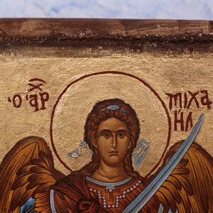 Puede incluir: Primer plano de un icono religioso que representa una figura con un halo, sosteniendo una espada. La figura tiene cabello casta&ntilde;o y viste una prenda roja y dorada. El fondo es dorado con texto rojo.