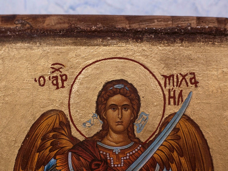 Puede incluir: Primer plano de un icono religioso que representa una figura con un halo, sosteniendo una espada. La figura tiene cabello casta&ntilde;o y viste una prenda roja y dorada. El fondo es dorado con texto rojo.