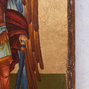 Puede incluir: Primer plano de un icono religioso que representa una figura con una capa roja, ropa azul y alas marrones. El icono tiene un fondo dorado con grietas y un marco de madera marr&oacute;n oscuro.