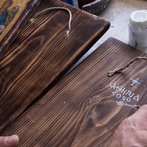 Puede incluir: Dos placas de madera con tinte marr&oacute;n oscuro. Una placa presenta un icono religioso con detalles dorados. La otra placa tiene texto blanco que dice "&Alpha;&Theta;&Eta;&Nu;&Alpha; 2020". Una mano est&aacute; pintando el texto en la segunda placa.