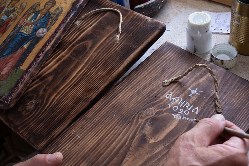 Puede incluir: Dos placas de madera con tinte marr&oacute;n oscuro. Una placa presenta un icono religioso con detalles dorados. La otra placa tiene texto blanco que dice "&Alpha;&Theta;&Eta;&Nu;&Alpha; 2020". Una mano est&aacute; pintando el texto en la segunda placa.