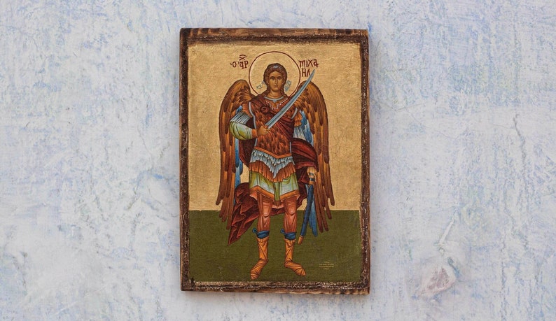 Puede incluir: Icono rectangular de madera que representa a un &aacute;ngel con una espada. El &aacute;ngel tiene grandes alas y viste una armadura. El fondo es dorado con texto en la parte superior. El marco es de color marr&oacute;n oscuro, con una pintura detallada.