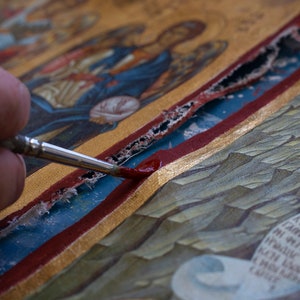 Puede incluir: Primer plano de un icono que se restaura con un pincel fino. El icono presenta pan de oro, pintura roja y azul, y figuras detalladas. El pincel aplica pintura roja a una zona da&ntilde;ada, revelando el proceso.