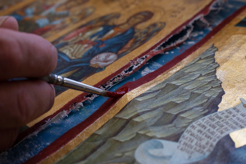 Puede incluir: Primer plano de un icono que se restaura con un pincel fino. El icono presenta pan de oro, pintura roja y azul, y figuras detalladas. El pincel aplica pintura roja a una zona da&ntilde;ada, revelando el proceso.