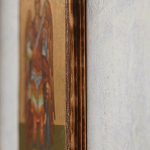 Puede incluir: Icono religioso que representa una figura alada vestida con ropas rojas y azules, sosteniendo una espada. La obra de arte est&aacute; sobre un fondo dorado y enmarcada por un borde de madera oscura y texturizada.