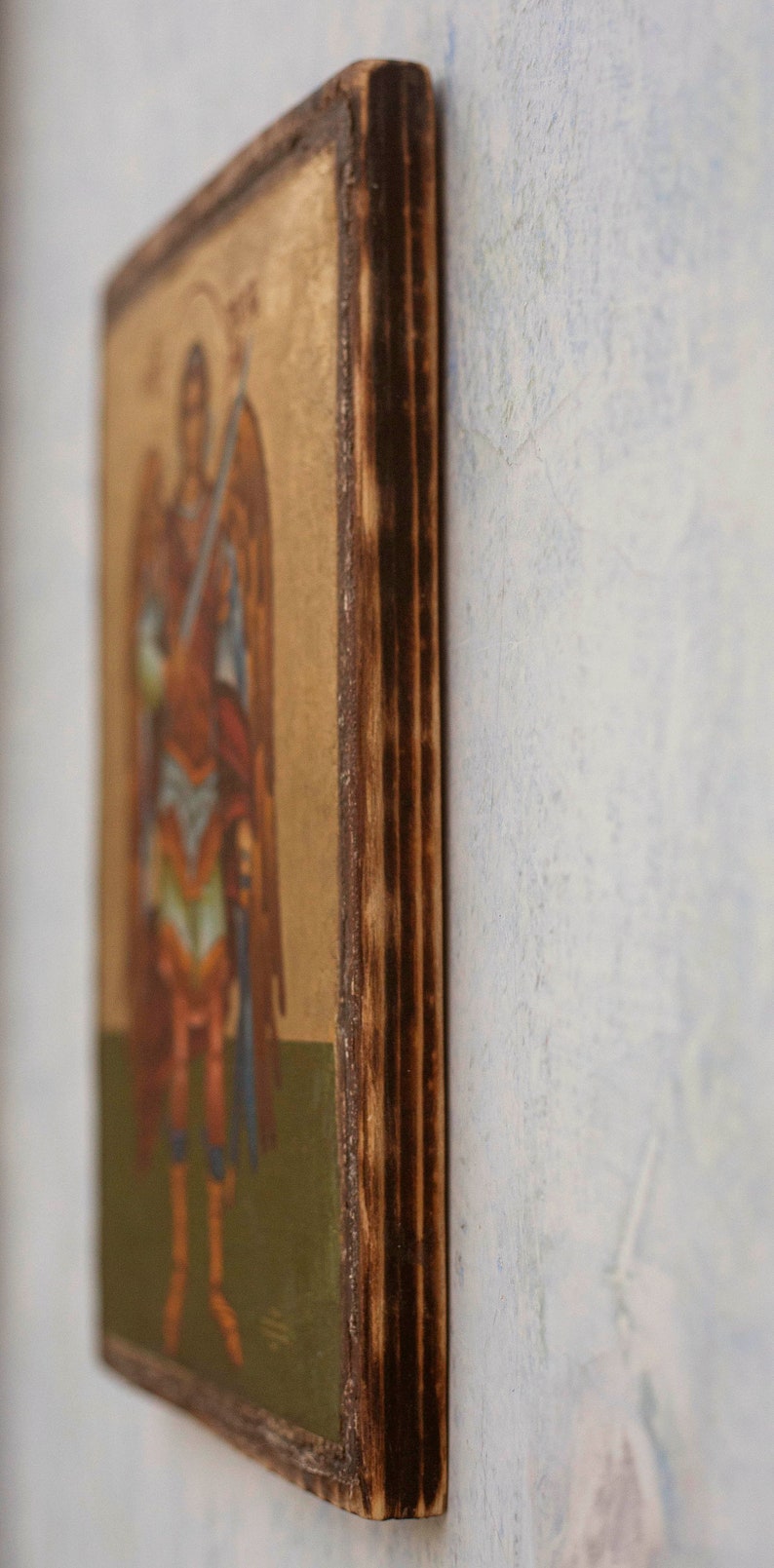 Puede incluir: Icono religioso que representa una figura alada vestida con ropas rojas y azules, sosteniendo una espada. La obra de arte est&aacute; sobre un fondo dorado y enmarcada por un borde de madera oscura y texturizada.