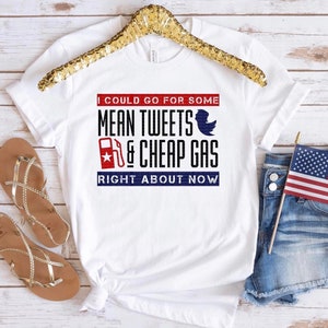 May include: White t-shirt with the text "I COULD GO FOR SOME MEAN TWEETS & CHEAP GAS RIGHT ABOUT NOW" in red and blue. The shirt features a gas pump graphic and a blue bird graphic. The shirt is displayed with sandals and denim shorts.