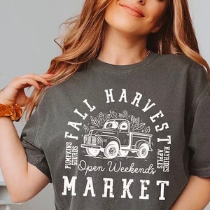 Może przedstawiać: Ciemnoszara koszulka z białym nadrukiem przedstawiającym zabytkową ciężarówkę i napisem "Fall Harvest Market". Projekt zawiera tekst dla "Pumpkins, Gourds, Hayrides, Apples" i "Open Weekends."