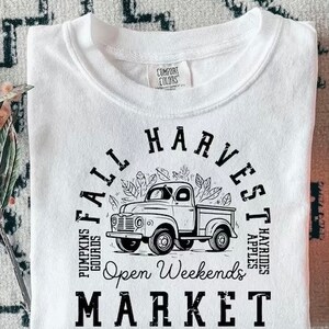 May include: White t-shirt with a black graphic of a vintage truck surrounded by leaves, with the words "Fall Harvest Market" and "Open Weekends". The shirt also says "Pumpkins Gourds" and "Hayrides Apples".