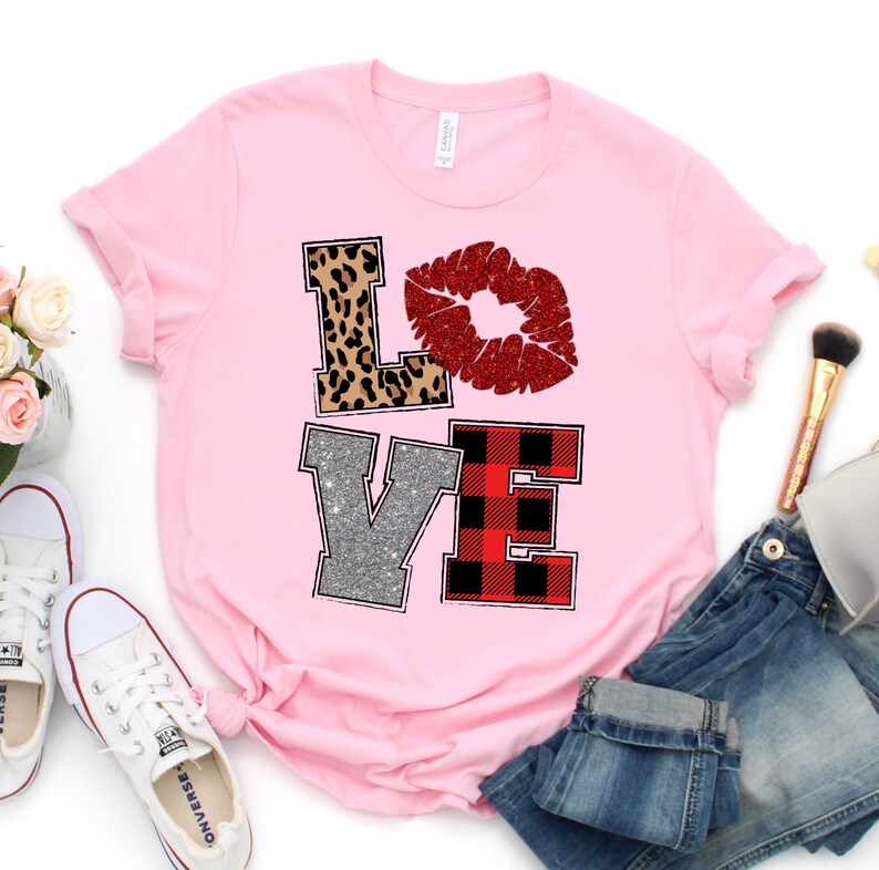 May include: A light pink t-shirt with the word "LOVE" in a decorative design. The "L" is leopard print, the "O" is a red glitter lip, the "V" is silver glitter, and the "E" is red and black plaid. The shirt is paired with jeans and white sneakers.