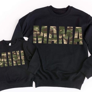Könnte beinhalten: Zwei schwarze Sweatshirts, eine Erwachsenen- und eine Kindergröße, mit den Wörtern "MAMA" und "MINI" im Camouflage-Druck auf der Brust. Die Sweatshirts werden auf weißem Hintergrund präsentiert.